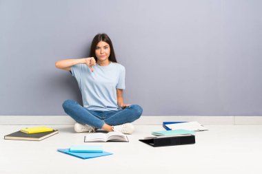 Yerde bir sürü kitabı olan genç bir bayan öğrenci baş parmağını aşağı gösteriyor.