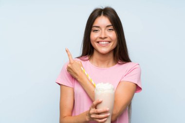 İzole edilmiş mavi arka planda çilekli milkshake 'i olan genç bir kadın bir ürünü sunmak için kenarı işaret ediyor.