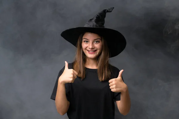 女の子とともに魔女衣装のためにハロウィンパーティー上の隔離された暗い背景与えますA親指アップジェスチャー — ストック写真