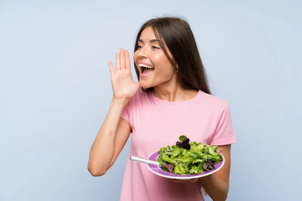 Young woman with salad over isolated blue background shouting with mouth wide open