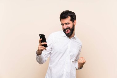 Sakallı genç adam cep telefonunu zafer pozisyonunda tutuyor.
