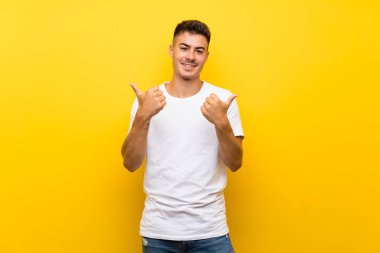 Soyutlanmış sarı arka planda genç yakışıklı bir adam. Baş parmakları havada ve gülümsüyor.