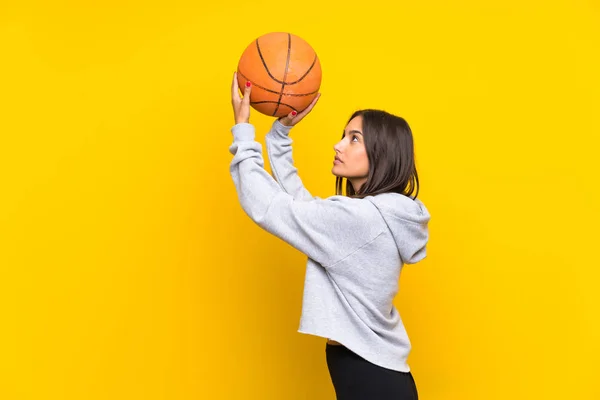 Young woman playing basketball over isolated yellow background