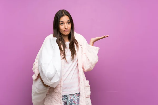 Young woman in pajamas and dressing gown over isolated purple background having doubts with confuse face expression