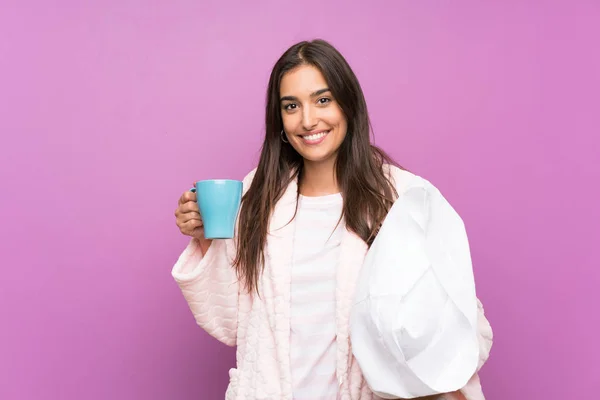 Mujer joven en pijama y bata sobre fondo púrpura aislado sosteniendo una taza de café — Foto de Stock