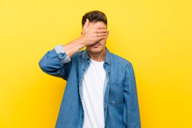 Elleri ile gözleri kaplayan izole sarı arka plan üzerinde genç yakışıklı adam