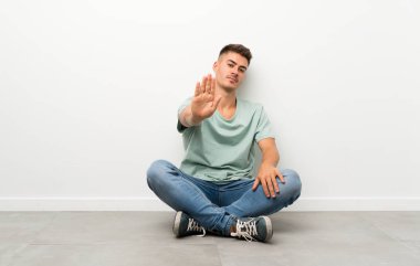 Genç yakışıklı adam yerde oturuyor ve eliyle jesti durduruyor.