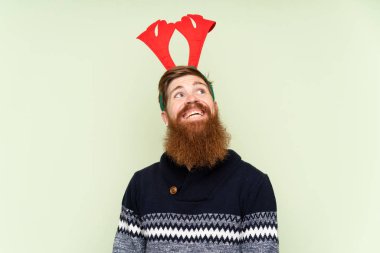 Kızıl saçlı, uzun sakallı, Noel tatili için giyinmiş yeşil arka planda saklanan ve gülümseyen bir adam.