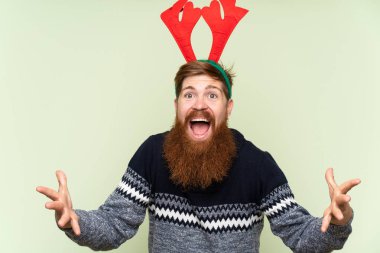 Kızıl saçlı, uzun sakallı, Noel tatili için giyinmiş yeşil arka planda, şok olmuş bir yüz ifadesi olan bir adam.