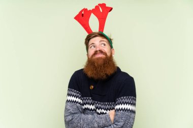 Kızıl saçlı, uzun sakallı bir adam, izole edilmiş yeşil arka planda Noel tatili için giyinmiş. Omuzlarını kaldırırken şüpheli hareketler yapıyor.