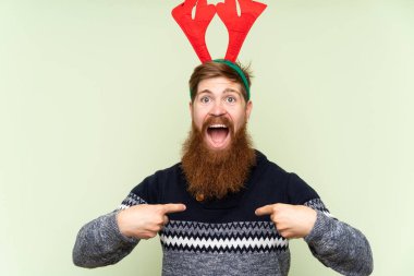 Kızıl saçlı, uzun sakallı, Noel tatili için giyinmiş, izole edilmiş yeşil arka planda, sürpriz bir yüz ifadesi olan bir adam.