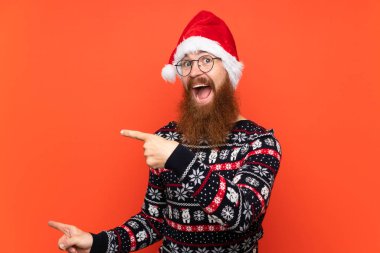 Uzun sakallı, soyutlanmış kırmızı arka planlı Noel adamı şaşırmış ve işaret ediyor.