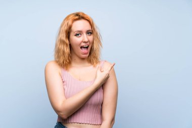İzole edilmiş mavi arka planı olan genç bir kadın şaşırmış ve işaret ediyor.