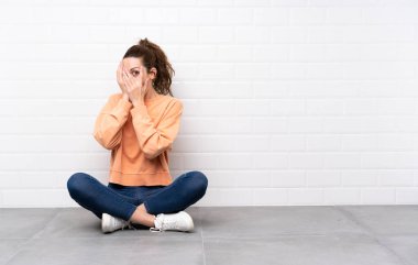 Kıvırcık saçlı genç bir kadın yerde oturuyor gözleri örtüyor ve parmaklarından bakıyor.