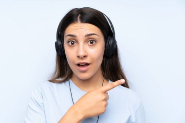 İzole edilmiş mavi arka planda müzik dinleyen genç bir kadın şaşırmış ve işaret ediyor.