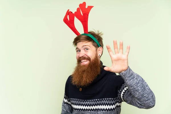 Redhead man with long beard dressed up for christmas holidays over isolated green background saluting with hand with happy expression