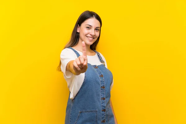 Jovem Mulher Dungarees Sobre Isolado Amarelo Fundo Mostrando Levantando Dedo — Fotografia de Stock