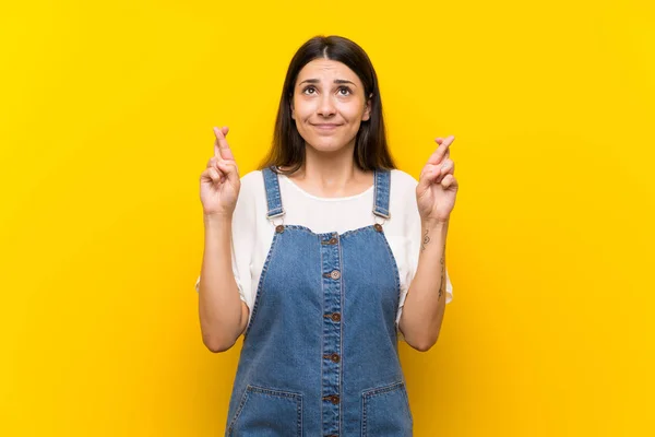 Jovem Mulher Dungarees Sobre Fundo Amarelo Isolado Com Dedos Cruzando — Fotografia de Stock