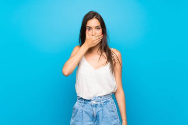 İzole edilmiş mavi arka planı olan genç bir kadın elleriyle ağzını kapatıyor.