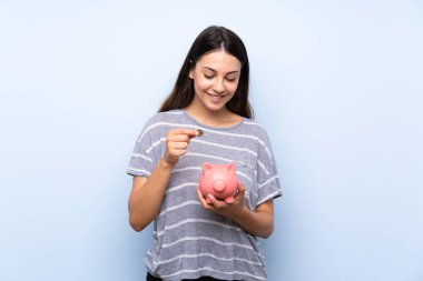 Büyük bir kumbara tutan izole mavi arka plan üzerinde genç esmer kadın