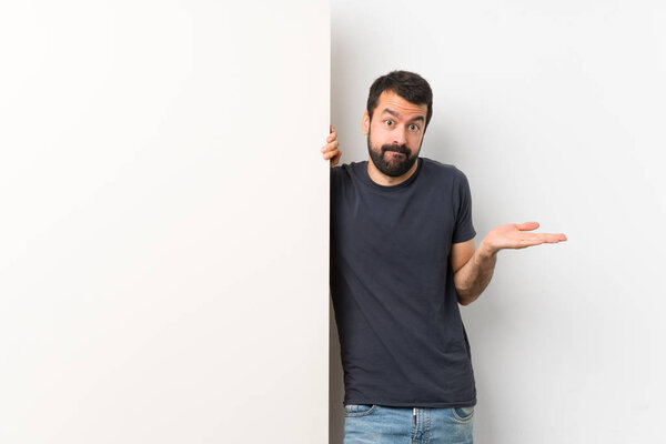 Young handsome man with beard holding a big empty placard having doubts while raising hands