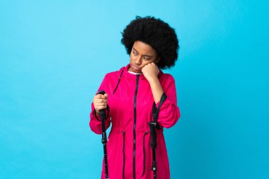 Sırt çantalı ve yürüyüş direkleriyle genç Afrikalı Amerikalı yorgun ve sıkılmış bir ifadeyle mavi arka planda izole edildi