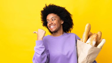 Genç Afrikalı Amerikalı bir kadın sarı arka planda izole edilmiş bir şey alıyor. Ürünü sunmak için yan tarafı işaret ediyor.