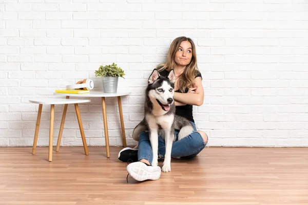 Young pretty woman with her husky dog sitting in the floor at indoors making doubts gesture while lifting the shoulders