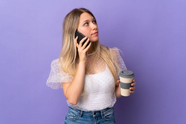 Mor arka planda izole edilmiş genç sarışın kadın elinde kahve ve bir cep telefonu tutuyor.