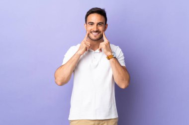 Mor arka planda yalnız, mutlu ve hoş bir ifadeyle gülümseyen genç yakışıklı bir adam.