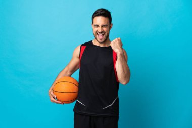 Genç yakışıklı adam mavi arka planda izole edilmiş basketbol oynuyor.