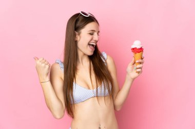 Pembe arka planda izole edilmiş kornet dondurması tutan mayo giymiş beyaz bir kadın zaferi kutluyor.