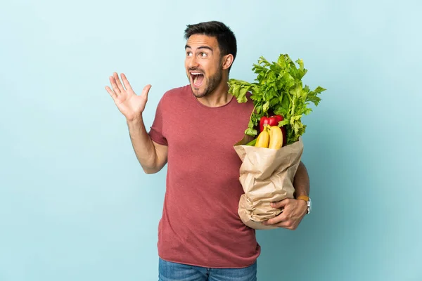 Młody Biały Człowiek Zakup Niektóre Warzywa Odizolowane Niebieskim Tle Zaskoczenie — Zdjęcie stockowe