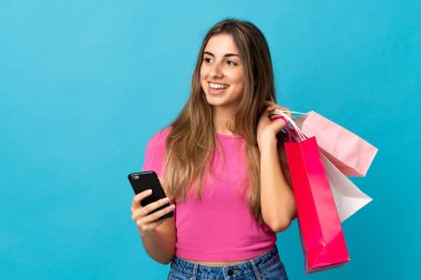 İzole edilmiş mavi arka planı olan genç bir kadın elinde alışveriş çantaları ve bir cep telefonu tutuyor.