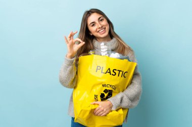 Geri dönüşüm için plastik şişelerle dolu bir çanta tutarken izole edilmiş mavinin üzerinde parmaklarıyla 