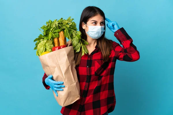 Young caucasian with vegetables and mask isolated on blue background having doubts and with confuse face expression