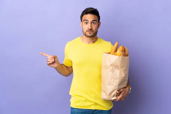 Young caucasian man holding a bag full of breads isolated on purple background pointing to the laterals having doubts