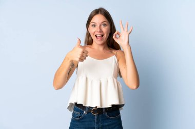 Genç Litvanyalı bir kadın mavi arka planda izole edilmiş. Tamam işareti ve baş parmak hareketi gösteriyor.