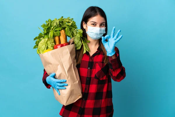 Young caucasian with vegetables and mask isolated on blue background showing ok sign with fingers