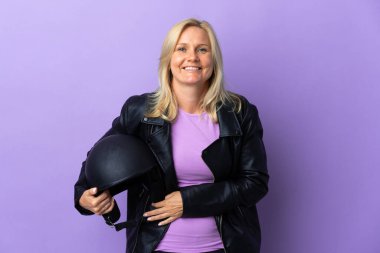 Mor arka planda izole edilmiş motosiklet kaskı tutan orta yaşlı kadın çok gülümsüyor.