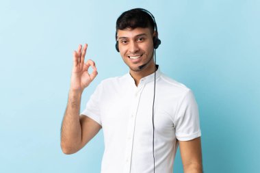 Kolombiyalı tele pazarlamacı izole edilmiş arka planda kulaklıkla çalışıyor. Parmaklarıyla tamam işareti gösteriyor.