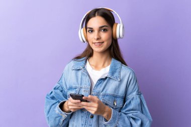 Mor arka planda izole edilmiş genç beyaz bir kadın cep telefonuyla müzik dinliyor ve ön tarafa bakıyor.