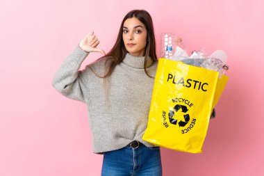 Pembe arka planda izole edilmiş plastik şişelerle dolu bir çanta taşıyan beyaz bir kadın, olumsuz bir ifadeyle baş parmağını gösteriyor.