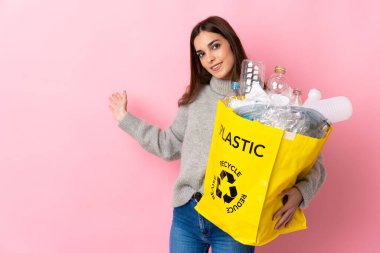 Pembe arka planda izole edilmiş plastik şişelerle dolu bir çanta taşıyan beyaz bir kadın davet edilmek için ellerini yana uzatıyor.