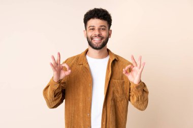 Faslı genç bir adam bej arka planda izole edilmiş. İki eliyle tamam işareti gösteriyor.