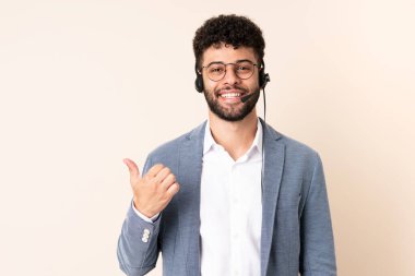 Faslı bir pazarlamacı bej arka planda izole edilmiş bir kulaklıkla çalışıyor. Ürünü sunmak için yan tarafı işaret ediyor.