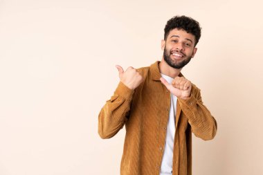 Bej arka planda izole edilmiş genç Faslı bir adam bir ürün sunmak için kenarı işaret ediyor.