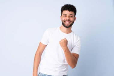 Genç Faslı bir adam mavi arka planda izole edilmiş zaferi kutluyor.