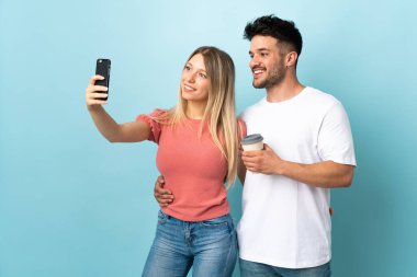 Mavi arka planda izole edilmiş genç beyaz bir çift cep telefonuyla selfie çekiyor.