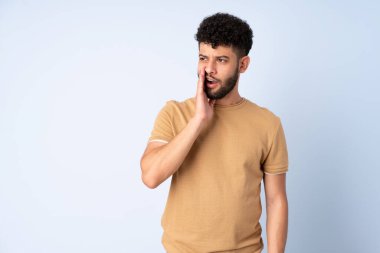 Faslı genç bir adam mavi arka planda izole edilmiş. Yan tarafa bakarken sürpriz bir hareketle bir şeyler fısıldıyor.